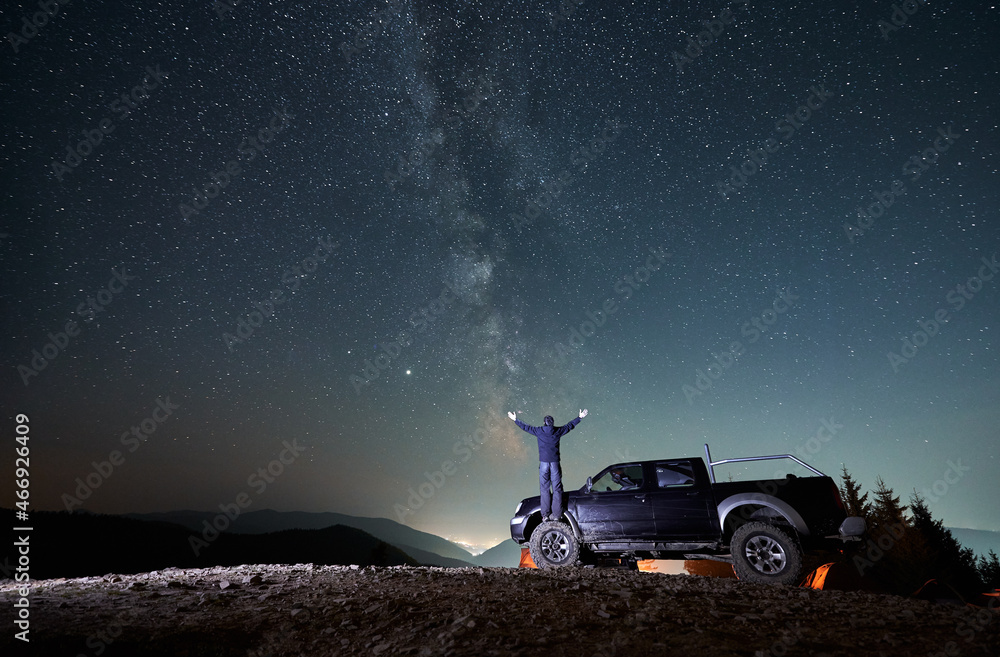 Back view of man with outstretched arms standing on rubber front wheel of black jeep in the middle of rocky mountain road and watching on night fairytale starry sky. The Milky Way over mountains. - obrazy, fototapety, plakaty 