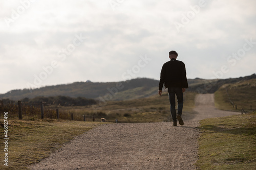 Man walking on an gravel path which leads to dunes, cloudy sky in the background