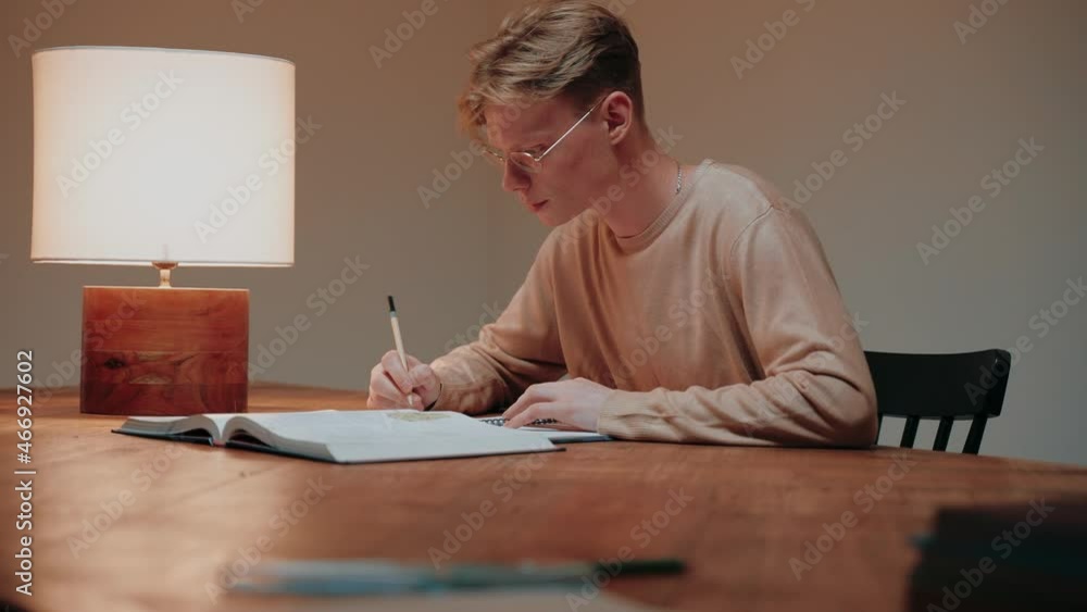 Young man studying at home writing in his notes, reading a textbook at the desk. Guy studying in warm bright room. Doing homework in the evening. High quality 4k footage Young man studying at home writing in his notes, reading a textbook at the desk. Guy studying in warm bright room. Doing homework in the evening. High quality 4k footage