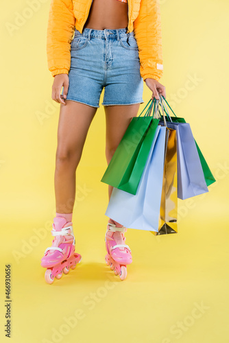 cropped view of african american young woman on pink roller skates holding shopping bags on yellow