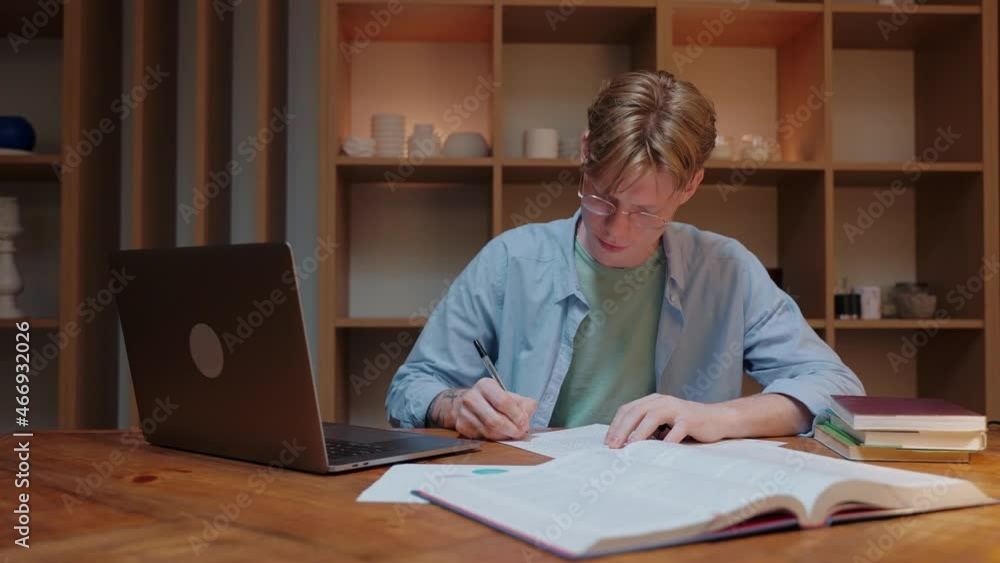 A young man studying at home looks at a laptop, writes a notebook and reads a textbook. Guy studying in warm wood room. Doing homework in the evening. High quality 4k footage A young man studying at home looks at a laptop, writes a notebook and reads a textbook. Guy studying in warm wood room. Doing homework in the evening. High quality 4k footage