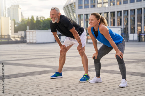 Sportive middle aged couple, man and woman looking away, taking a short break while jogging together in the city on a warm summer day