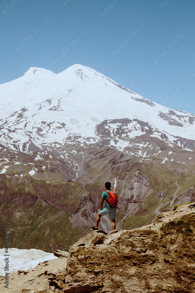Man with red backpack stand on rocky mountain and look at snowy peak of Elbrus.