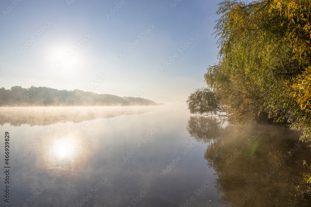 Autumn landscape in the early morning on the river. Misty water surface. Yellow leaves on trees and bushes are illuminated by the rays of the rising sun. Dawn on a cold autumn morning.