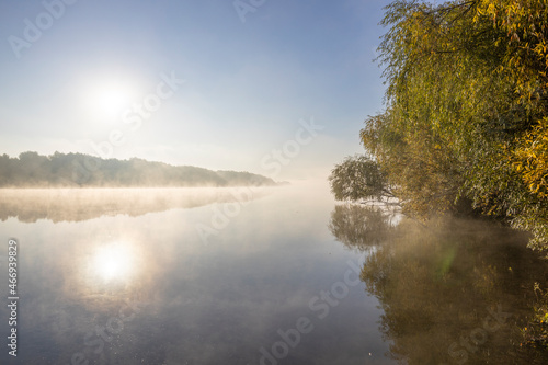 Autumn landscape in the early morning on the river. Misty water surface. Yellow leaves on trees and bushes are illuminated by the rays of the rising sun. Dawn on a cold autumn morning.