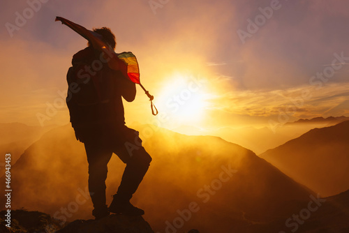 backlit silhouette of hiker with backpack carrying a rainbow LGBT pride flag on top of a mountain watching the sunset. sport and healthy life concept.