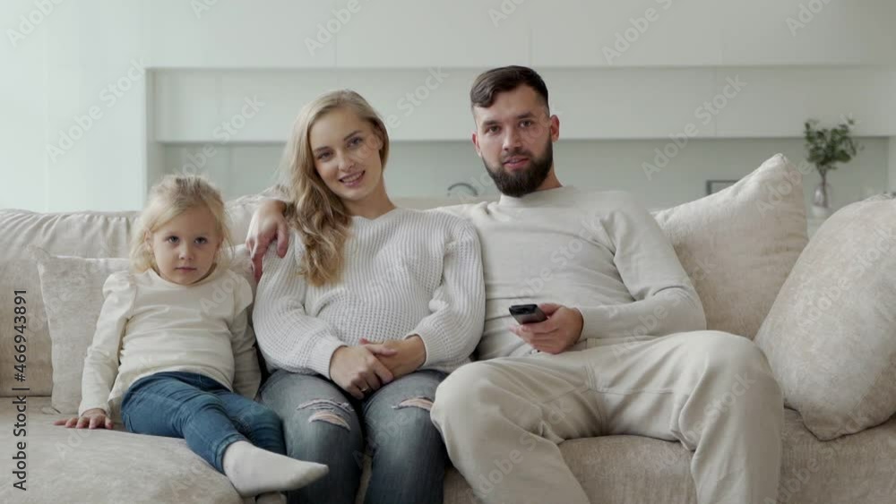 Young parents and their cute little daughter are watching an interesting informative documentary at home on the couch Young parents and their cute little daughter are watching an interesting informative documentary at home on the couch