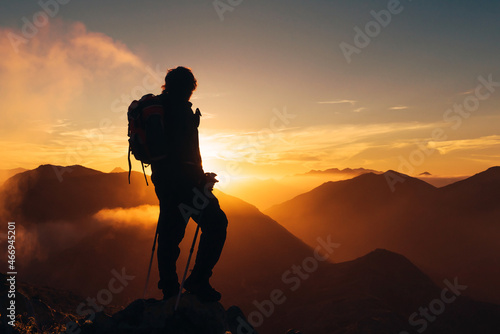 backlit silhouette of hiker with backpack and poles on top of a mountain watching the sunset. sport and healthy life concept.