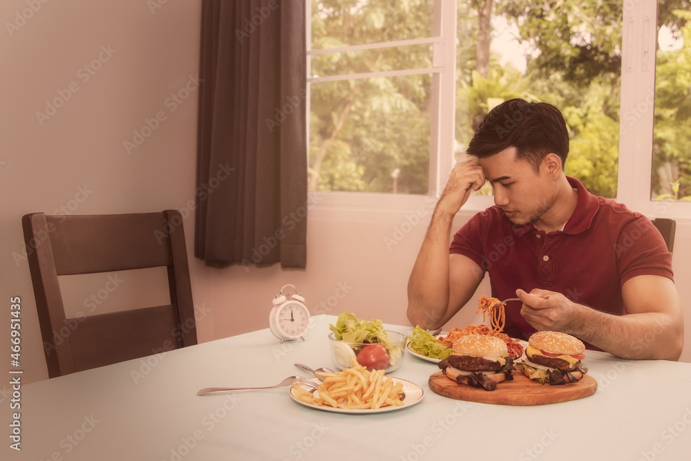 Concept of reducing junk food for health : handsome guy wants to have lunch, but the table is full of junk food such as french fries, hamburgers, and he pondered to control the food for good health. - obrazy, fototapety, plakaty 