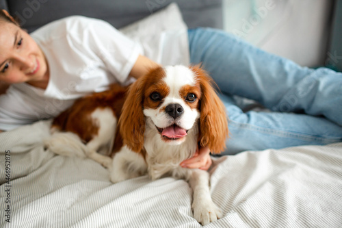 Young woman lying on the bed at home with Cavalier King Charles Spaniel dog and smiling.