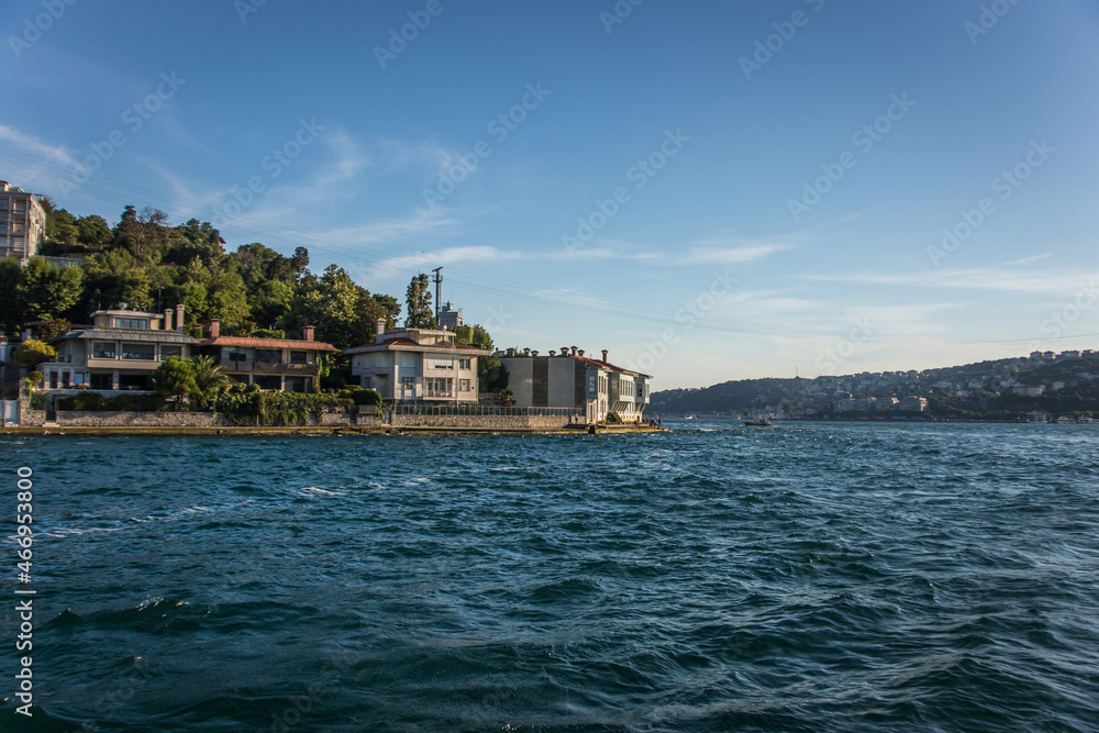 Luxurious houses and mansions over the hills located on the shore of Istanbul Bosphorus strait with green trees on the back and blue water in front of the houses.