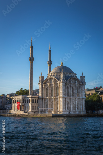 Exterior of the famous Ortakoy Mosque or Grand Imperial Mosque in Istanbul, Turkey under the Bosphorus Bridge with a Turkish flag hanging on one of it's walls under the blue sky.