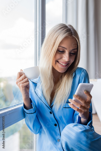happy young woman holding smartphone and cup of coffee near window