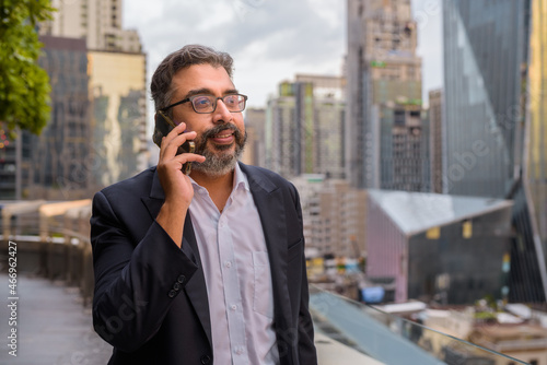 Portrait of handsome Indian businessman in city at rooftop garden talking on phone