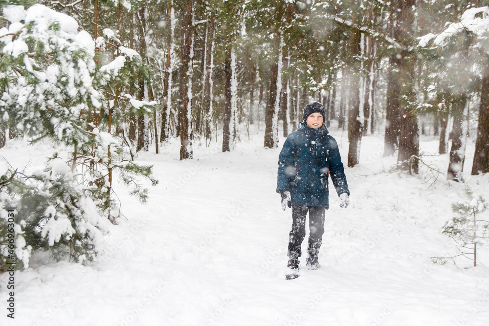 Front view of child boy in winter forest in snowstorm. Kid on snowy winter background. Actively spending time outdoors. Winter snowy woodland. Cold frost weather in snowstorm. Happy childhood