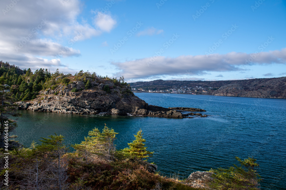 A rugged coastline in the Atlantic Ocean. The rugged cliffs of rocks have some vegetation and trees. It's a bright sunny day with a blue sky and few clouds. The ocean is deep blue with some waves.