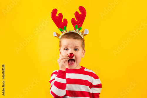 Happy funny toddler boy in red nose and horns reindeer with gift on yellow colored background