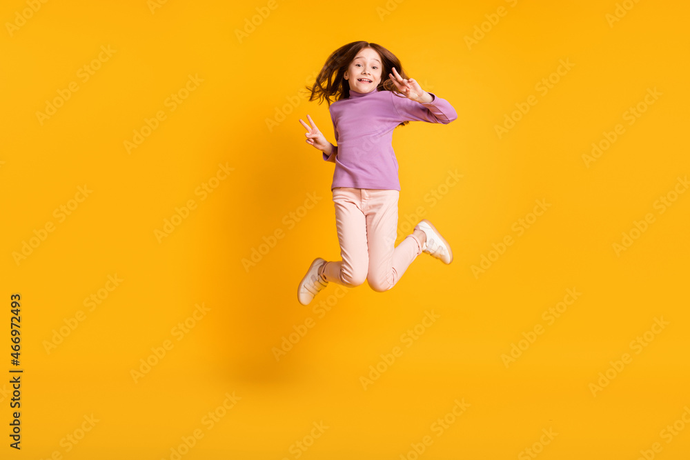Full size photo of happy excited positive smiling little girl jumping show v-sign isolated on yellow color background