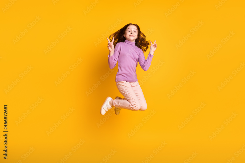 Full size photo of happy excited smiling cheerful little girl jumping showing v-sign isolated on yellow color background Full size photo of happy excited smiling cheerful little girl jumping showing v-sign isolated on yellow color background