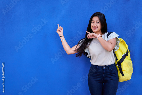 Young latina woman, slim college student, blue jeans, light blouse, with yellow backpack, on blue background, pointing to copy space.