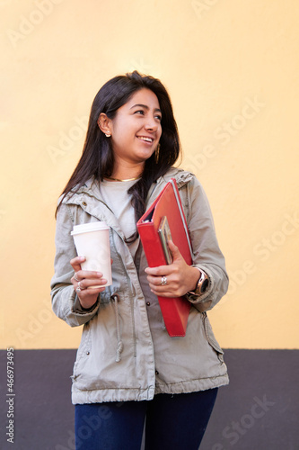 young, slim Latina female student, holding her notebook, folder in one hand and in the other her coffee, looking at copy space