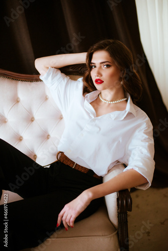 Attractive young girl with dark hair with Hollywood styling and makeup and red lipstick in a white men's shirt and dark trousers with a string of pearls around her neck in a classic interior.
