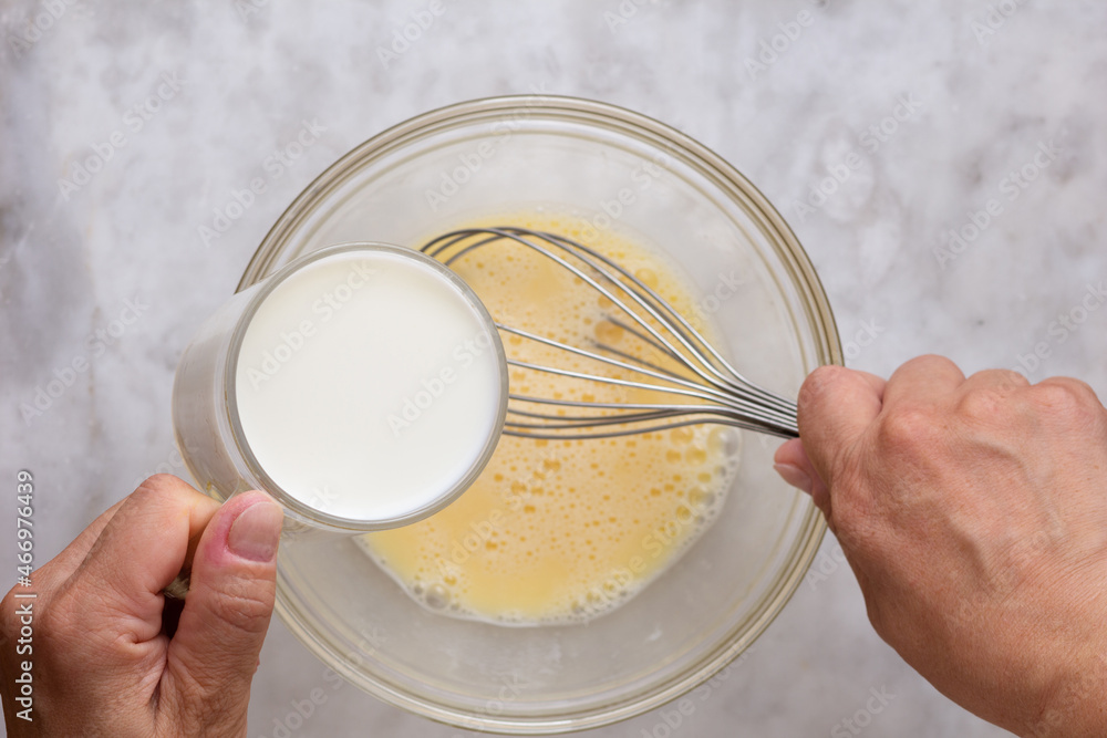 Top view of woman hand holding glass of milk putting into mixed raw eggs in glass bowl on marble surface Top view of woman hand holding glass of milk putting into mixed raw eggs in glass bowl on marble surface