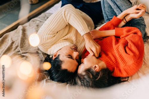 Passionate romantic couple spending home with garlands. Young woman and man are lying on bed in sweaters and hugging