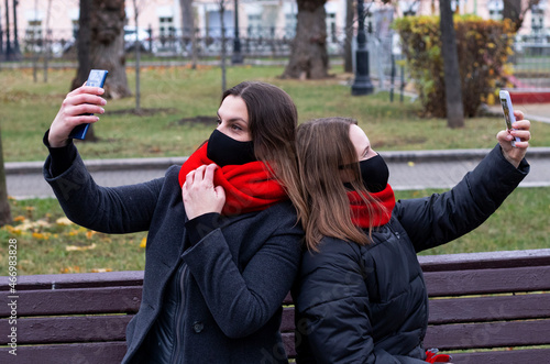 two girls friends sit on a bench in an autumn park and take a selfie on the phone