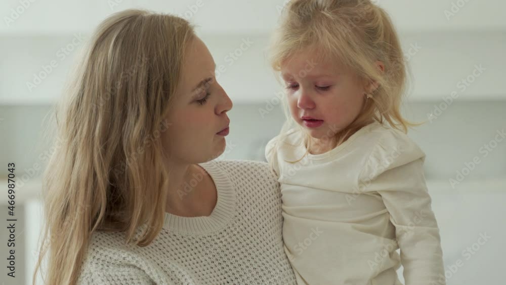 Child is crying in the room in the arms of his mother. Young mother tries to calm her crying little daughter. The concept of raising a child. Child is crying in the room in the arms of his mother. Young mother tries to calm her crying little daughter. The concept of raising a child.