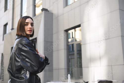 Successful self-confident attractive woman, business person, freelancer, entrepreneur, copywriter holding laptop in her hands and looking at camera against modern office corporate buildings background