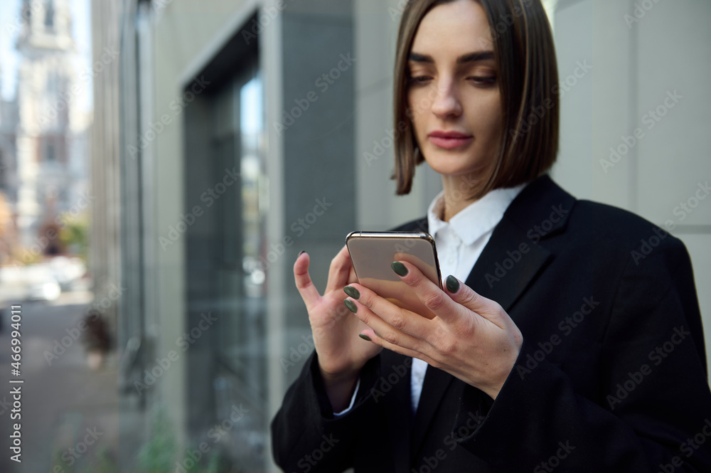Lifestyle business portrait of female freelancer, motivated young successful lady holding and swiping mobile phone against modern corporate building background. Business and communication concept