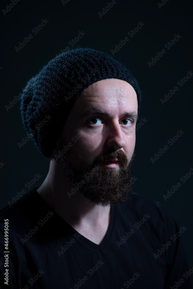 authentic dramatic portrait of a millennial with a beard in a black knitted hat, on a dark background authentic dramatic portrait of a millennial with a beard in a black knitted hat, on a dark background