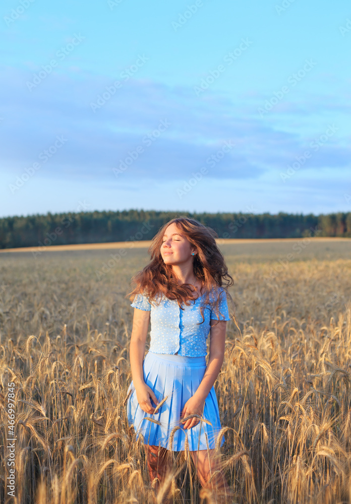 Happy teen girl is smiling at wheat field, touching ears of wheat with her hand. Beautiful teenager enjoying nature in warm sunshine in a wheat field on sunset background. Happy teen girl is smiling at wheat field, touching ears of wheat with her hand. Beautiful teenager enjoying nature in warm sunshine in a wheat field on sunset background.