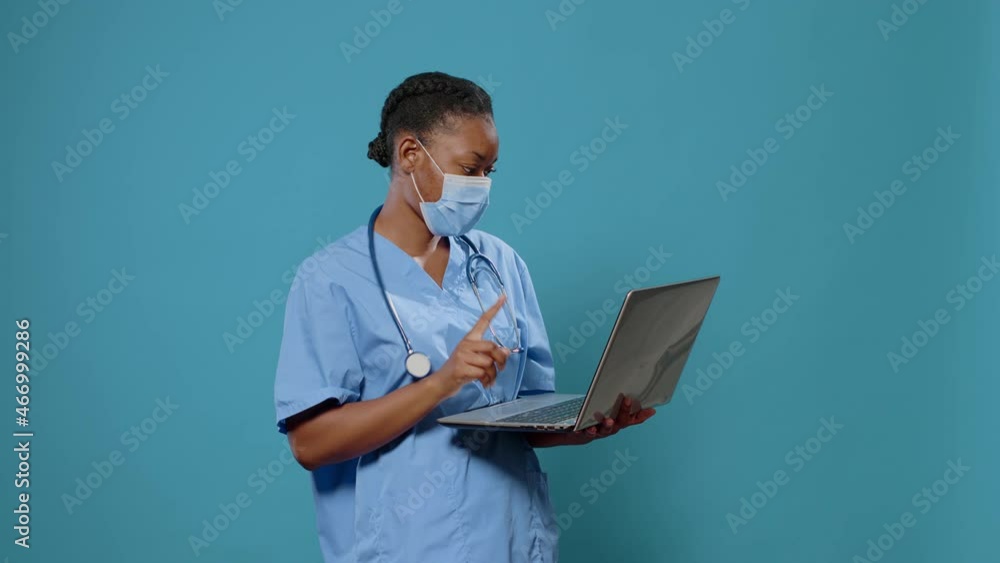 Woman nurse wearing face mask and using laptop in front of camera. Medical assistant in uniform having protection against coronavirus while holding computer to work on healthcare. Woman nurse wearing face mask and using laptop in front of camera. Medical assistant in uniform having protection against coronavirus while holding computer to work on healthcare.