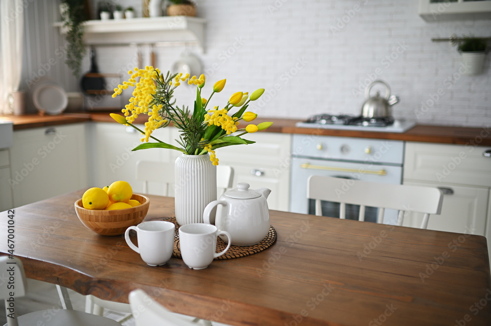 Obraz premium a bouquet of yellow tulips and mimosa in a vase on a wooden table against the background of a white modern kitchen, white dishes and lemons.