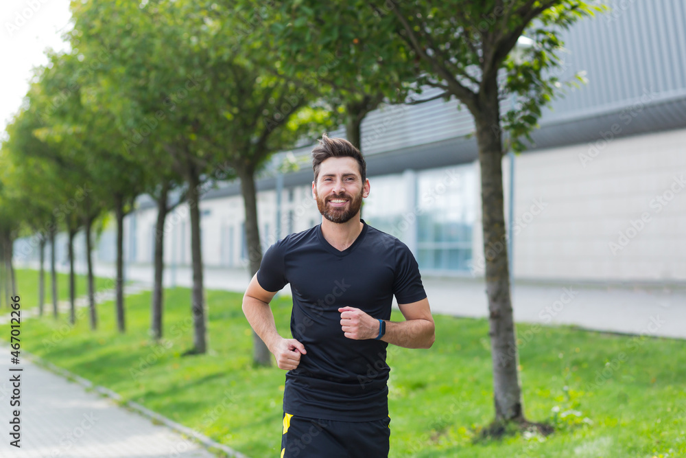 Young Caucasian bearded happy man runner jogging down the street on urban background of city park. Morning jog. Active healthy lifestyle fitness. Sport workout outdoor. Athlete training run exercise Young Caucasian bearded happy man runner jogging down the street on urban background of city park. Morning jog. Active healthy lifestyle fitness. Sport workout outdoor. Athlete training run exercise