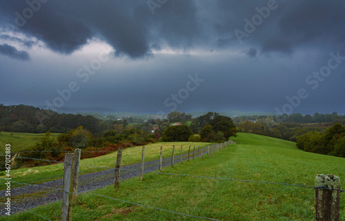 green grass field fenced with a fence of wooden sticks and barbed wire. Next to a dirt road and stones parallel to the fence in the background trees and the very dark sky with very gray clouds of rain