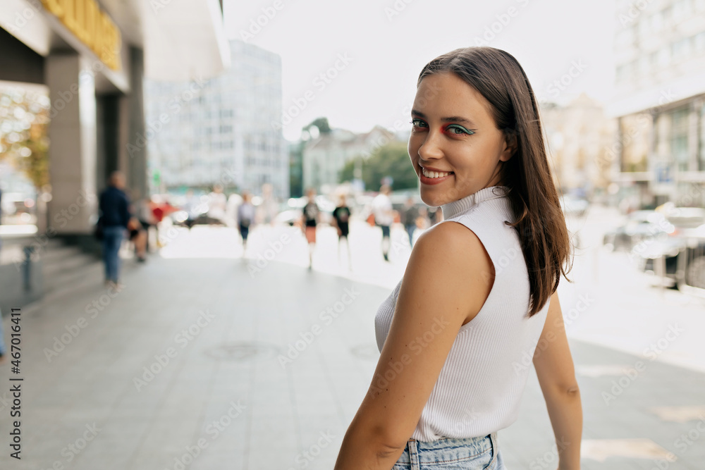 Close up portrait of adorable young lady with brunette hair wearing white t-shirt turn around at camera on city background