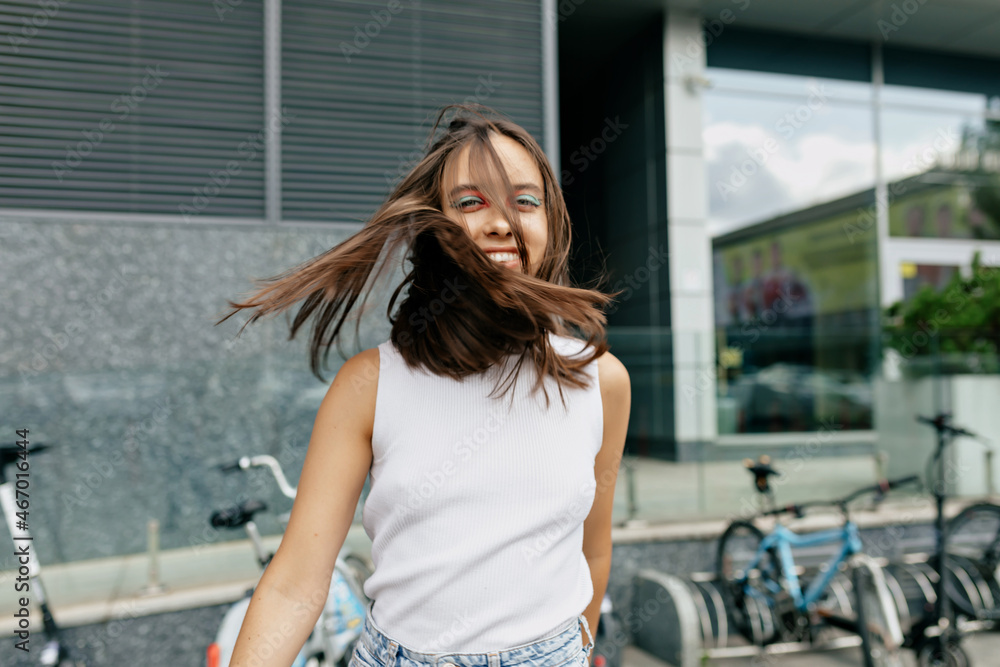 Fototapeta premium Excited charming woman with happy smile wearing white t-shirt is spinning around on the background of city