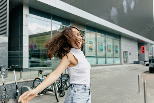 Attractive funny girl with flying dark hair looking at camera and spend time outside in good warm day in the city