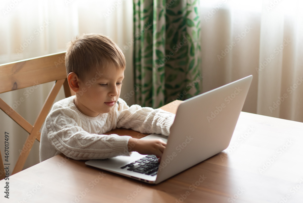 Little boy in a white sweater sits at a laptop and types a letter to Santa Claus. Distant education concept