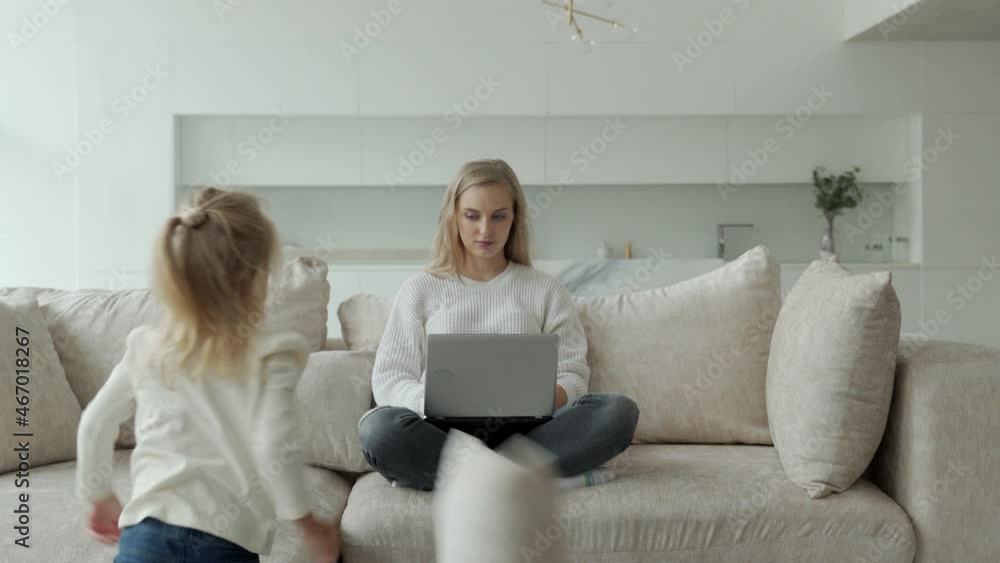 Female entrepreneur focused on working on a laptop on the couch while her daughter plays and prevents her mom from working Female entrepreneur focused on working on a laptop on the couch while her daughter plays and prevents her mom from working