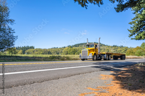 Bright yellow day cab big rig semi truck with empty flat bed semi trailer running on the narrow road to warehouse for the next load