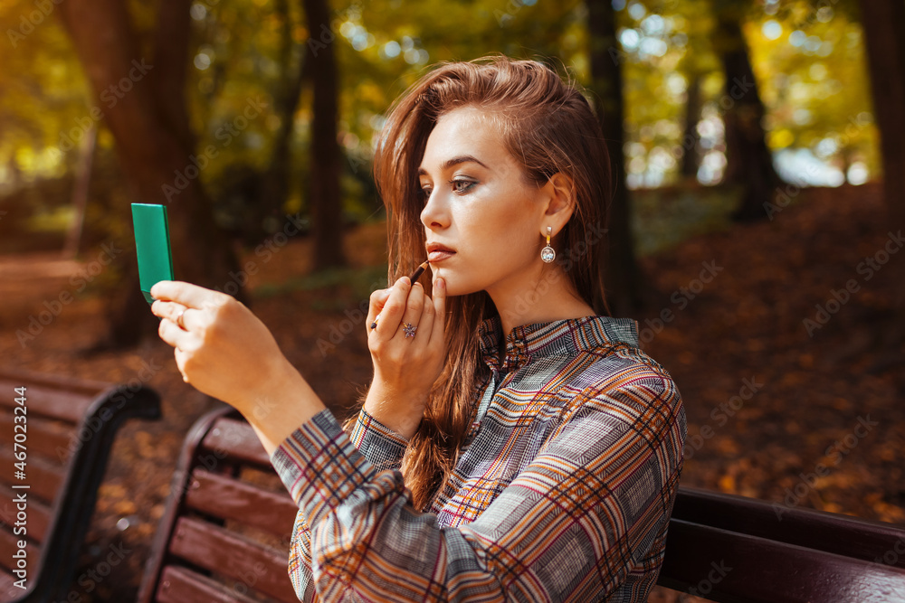 Portrait of woman applying lipstick using hand mirror in fall park sitting  on bench. Girl checking makeup outdoors Stock Photo | Adobe Stock