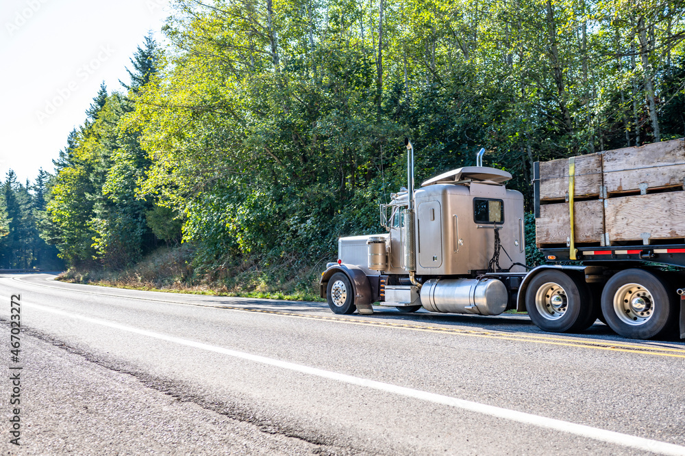 Classic big rig bonnet semi truck with roof spoiler transporting harvest in wood boxes fastened on flat bed semi trailer running for delivery on the highway road with trees on the hill