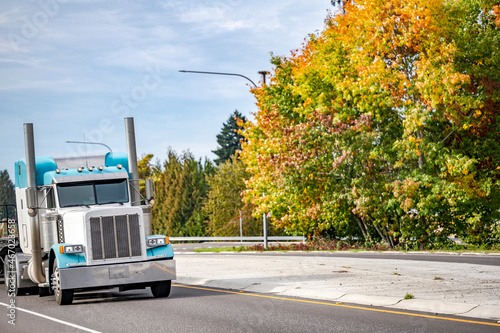 Classic bonnet white and blue big rig semi truck tractor transporting cargo on flat bed semi trailer running on the autumn road © vit