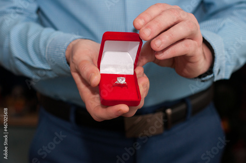 An open red velvet box with a white gold ring with a precious stone in the hands of a man in a blue shirt