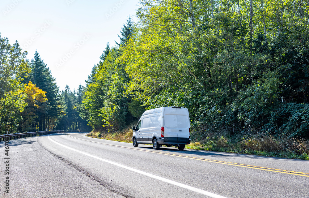 Compact cargo commercial mini van driving on the turning road with forest on the sides Compact cargo commercial mini van driving on the turning road with forest on the sides