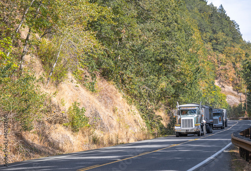 Convoy of the big rig tipper semi trucks with tip trailers running on the mountain winding road with trees on the hills